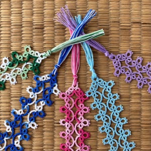 May include: A collection of handmade lace bookmarks in various colors, including green, blue, pink, and purple. Each bookmark features a detailed floral design and is topped with a matching tassel. The bookmarks are displayed on a woven mat.