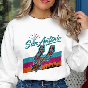 May include: A white crewneck sweatshirt featuring a teal embroidered San Antonio script logo above a stylized spur icon. The design includes horizontal stripes in teal, magenta, and orange, with a basketball and city skyline graphic.