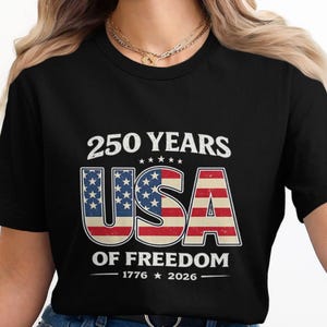 May include: Black t-shirt with a patriotic design celebrating 250 years of American freedom. The design features the text "250 YEARS USA OF FREEDOM 1776-2026" in white, red, and blue, with the USA letters incorporating the American flag.