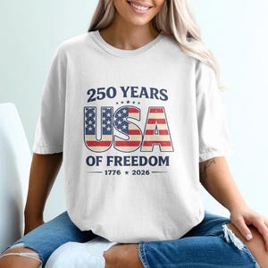 May include: White t-shirt with a patriotic design celebrating 250 years of American freedom. The design features the text "250 YEARS USA OF FREEDOM" with the USA letters in a flag-inspired design. The shirt includes the years 1776 and 2026.