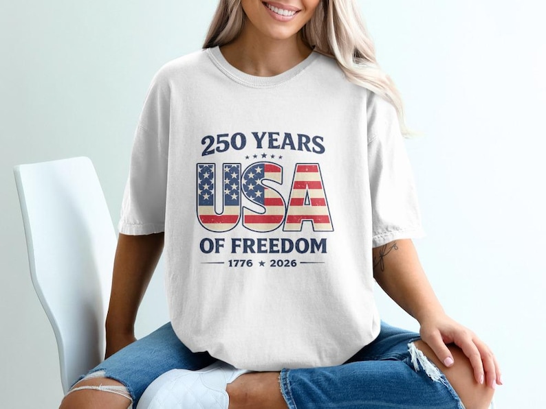 May include: White t-shirt with a patriotic design celebrating 250 years of American freedom. The design features the text "250 YEARS USA OF FREEDOM" with the USA letters in a flag-inspired design. The shirt includes the years 1776 and 2026.