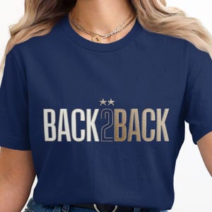 Peut inclure: Un t-shirt bleu marine à manches courtes avec le texte BACK 2 BACK en lettres blanches et dorées. Deux petites étoiles dorées sont placées au-dessus du chiffre 2. Le t-shirt présente une encolure ronde et une coupe standard avec un motif.