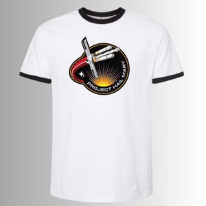 Puede incluir: Camiseta blanca con ribete negro en el cuello y las mangas. La camiseta presenta un gráfico circular con el texto "PROJECT HAIL MARY" y un diseño de una nave espacial y un sol.