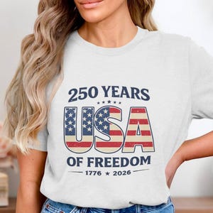 May include: White t-shirt with a patriotic design celebrating 250 years of American freedom. The design features the text "250 YEARS USA OF FREEDOM 1776-2026" in navy blue and red, white, and blue colors.