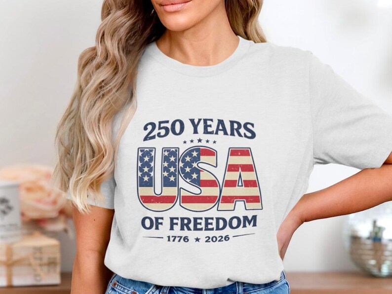 May include: White t-shirt with a patriotic design celebrating 250 years of American freedom. The design features the text "250 YEARS USA OF FREEDOM 1776-2026" in navy blue and red, white, and blue colors.