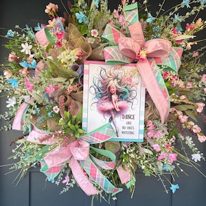 May include: A decorative wreath with a variety of faux flowers in pink, blue, and white. The wreath features a sign with a ballerina illustration and the text "DANCE LIKE NO ONE'S WATCHING". Pink and green plaid ribbons and bows are also included.