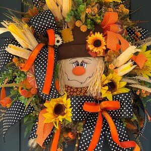 May include: A fall-themed wreath featuring a scarecrow face with a brown hat, sunflowers, and corn. The wreath is decorated with black and white polka dot ribbon and orange ribbon. The wreath is a festive decoration for the autumn season.