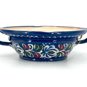 May include: A deep blue ceramic bowl featuring two side handles and a hand-painted floral pattern in white, green, and red. The interior of the bowl is a light cream color, and the exterior displays a decorative scalloped border near the rim.