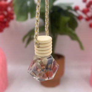 May include: A clear glass bottle with a geometric shape filled with colorful stones and a wooden cap. The bottle is hanging from a beige and gold cord.