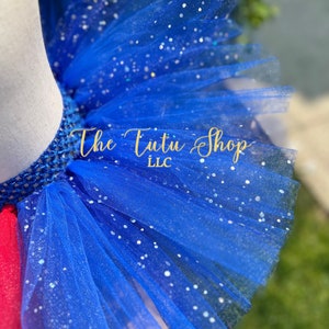 Może przedstawiać: Tutu z niebieskiego tiulu z srebrnym brokatem. Tutu ma haftowaną gumkę i widoczny jest napis "The Tutu Shop LLC" na tkaninie.