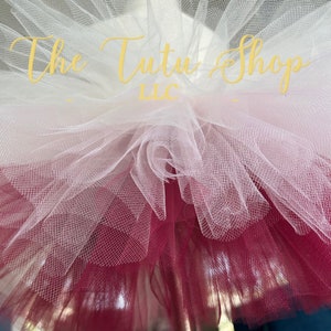Może przedstawiać: Tutu z tiulu w kolorze białym i bordowym z napisem "The Tutu Shop LLC" na górnej warstwie.