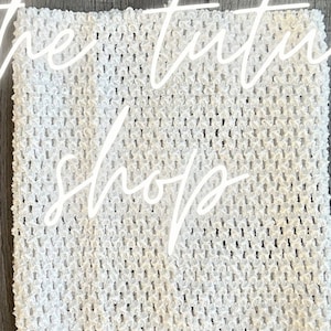 May include: Close-up of white, textured fabric with a knitted or crocheted appearance. The fabric has small holes throughout. The text "the tutu shop" is overlaid in a cursive font. The background is a neutral colour.