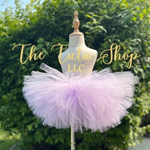 Może przedstawiać: Tutu z tiulu lawendowego na białym manekinie. Tutu jest pełne i puszyste, z wieloma warstwami tiulu. Tekst "The Tutu Shop LLC" jest widoczny w tle.