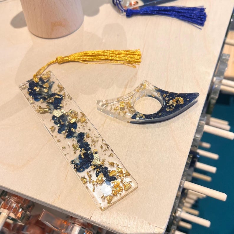 May include: A rectangular resin bookmark with a gold tassel and a crescent-shaped resin page holder, both featuring a clear base with embedded dark blue pigment swirls and gold leaf flakes. A separate blue tassel is visible at the top edge.
