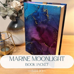 May include: A hardcover book featuring a dust jacket with an abstract alcohol ink design in shades of deep blue, purple, and magenta, accented with thin, shimmering gold veins. The book has a gold ribbon bookmark and a dark blue top edge.