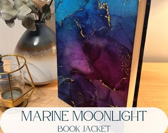 Puede incluir: Un libro de tapa dura con una sobrecubierta con un diseño abstracto de tinta alcohólica en tonos azul profundo, morado y magenta, acentuado con vetas doradas. El libro tiene un borde de página blanco y un marcapáginas de cinta dorada.