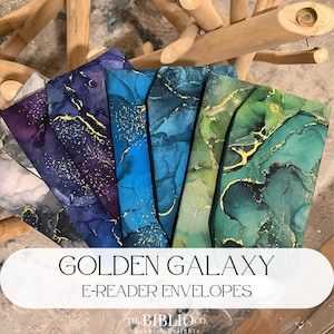 Op de afbeelding: Een collectie van vijf stoffen e-readerhoezen met abstracte marmerpatronen in tinten grijs, paars, blauw, groenblauw en groen, elk geaccentueerd met goudkleurige aders. De tekst GOLDEN GALAXY E-READER ENVELOPES staat eronder gedrukt.