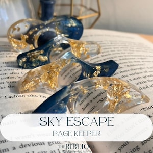 May include: A set of four resin page keepers featuring a deep blue and clear design with embedded gold leaf flakes. Each curved, thumb-hole accessory is positioned on an open book page. The text SKY ESCAPE PAGE KEEPER is printed on the image.