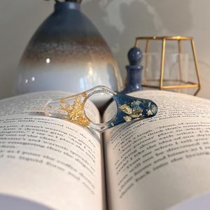 May include: A resin page holder with a central finger hole, featuring a clear section with embedded gold leaf on one side and a deep blue section with embedded gold leaf on the other, resting on the open pages of a printed book.