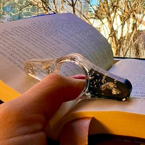 May include: A clear resin book page holder with a thumb hole, featuring embedded gold leaf flakes and a dark green section. The accessory is shaped like a rounded triangle and is positioned on an open book with printed text on the pages.