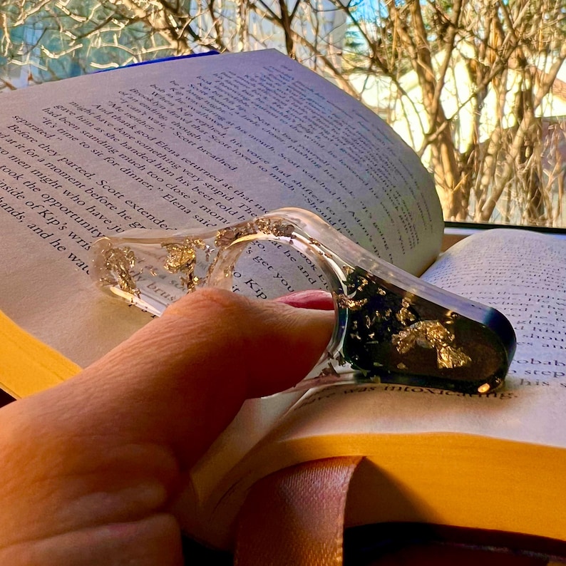 May include: A clear resin book page holder with a thumb hole, featuring embedded gold leaf flakes and a dark green section. The accessory is shaped like a rounded triangle and is positioned on an open book with printed text on the pages.