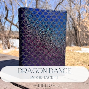 May include: A rectangular book jacket titled DRAGON DANCE BOOK JACKET by THE BIBLIO CO. bookish delights. The cover features a holographic, iridescent scale pattern that transitions from deep blue at the top to purple at the bottom.