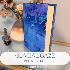 May include: A hardcover book featuring a book jacket with a vibrant blue, purple, and teal alcohol ink pattern accented by thin gold veins. The book has cream-colored pages and a thin yellow ribbon bookmark extending from the top edge.