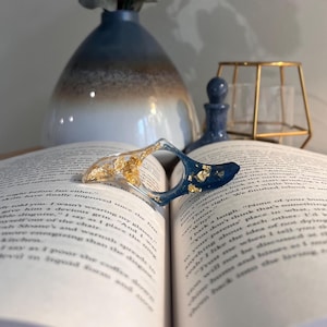 May include: A curved, crescent-shaped resin page holder resting on an open book. The object features a clear section filled with gold leaf flakes on one side and a deep navy blue section with gold leaf flakes on the other, joined in the center.