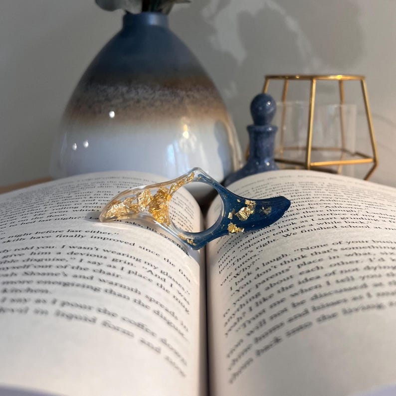 May include: A curved, crescent-shaped resin page holder resting on an open book. The object features a clear section filled with gold leaf flakes on one side and a deep navy blue section with gold leaf flakes on the other, joined in the center.