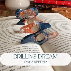May include: Three resin page holders with a dark blue and clear gradient finish, embedded with metallic copper flakes. Each tool features a central circular opening for a thumb, designed to keep book pages open while reading.
