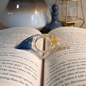 May include: A crescent-shaped page holder made of clear resin, featuring a deep blue gradient on one side and embedded gold leaf flakes throughout the transparent section. The object has a central circular opening for a thumb to rest while reading.
