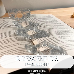 May include: Three clear resin page holders with embedded silver glitter flakes, each featuring a central circular cutout. These functional accessories are positioned on the open pages of a book, displaying the text IRIDESCENT IRIS PAGE KEEPER.