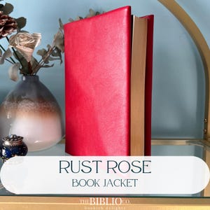 May include: A bright red book jacket with a textured finish, standing upright. The text on the overlay reads RUST ROSE BOOK JACKET and THE BIBLIO CO. bookish delights. The book has tan-colored pages visible along the side edge of the cover.