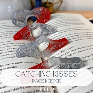 May include: A set of four resin page holders, each featuring a dual-tone design with one side in solid red and the other in translucent silver glitter. These thumb-hole book accessories are shaped like curved wings and rest on an open book page.