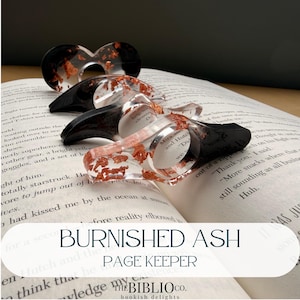 Puede incluir: Protectores de página de resina transparente con escamas de cobre y detalles negros descansan sobre un libro abierto. Los protectores de página tienen un diseño circular con un asa curvada. El texto "BURNISHED ASH PAGE KEEPER" es visible.