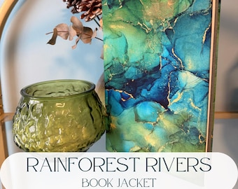 Puede incluir: Un libro de tapa dura con un diseño de cubierta abstracto y vibrante en tonos verde azulado, verde esmeralda y azul profundo con vetas doradas metálicas. Junto al libro hay un cuenco de cristal verde oliva texturizado y translúcido.