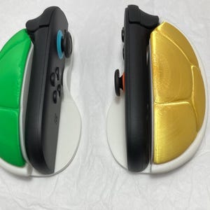 May include: Two black game controllers with green and gold shell-shaped grips. The controllers are set on white bases. The green grip has a blue joystick, and the gold grip has an orange joystick. The controllers are designed for gaming.