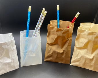 May include: Four paper bags, two white and two brown, stand upright. Pencils with blue, silver, and red barrels are visible inside the bags. The bags have a crumpled texture, suggesting they are made of paper.
