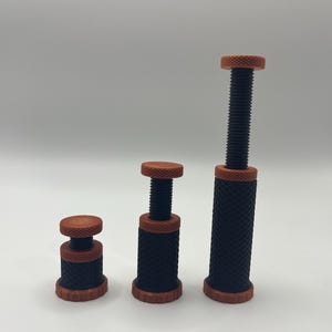 May include: Three adjustable screw jacks of varying heights, each featuring a black cylindrical base with a diamond-patterned knurled texture, orange circular top and bottom plates, and a central black threaded screw mechanism for height adjustment.