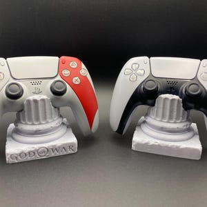 May include: Two white and gray game controllers, one with red accents, rest on matching gray stands. The stands are shaped like columns and have the words "GOD OF WAR" printed on them. The controllers are for a gaming console.
