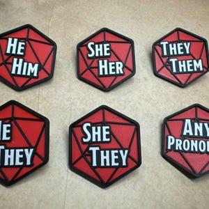 May include: Six red and black hexagonal pins with white text. The pins read "He Him", "She Her", "They Them", "He They", "She They", and "Any Pronouns".