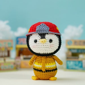 May include: A crocheted penguin wearing a red and gray firefighter hat and a yellow and black firefighter uniform.
