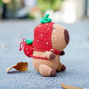 Może przedstawiać: Pluszowa zabawka wykonana na szydełku, nosząca czerwony kapelusz w kształcie truskawki z zielonymi liśćmi. Zabawka jest brązowa i ma małe, okrągłe ciało z długim pyskiem.