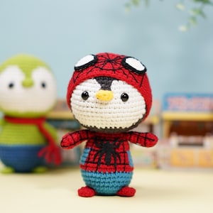 Puede incluir: Un pingüino de crochet con un disfraz de Spiderman rojo y negro. El pingüino está de pie sobre una superficie marrón claro.