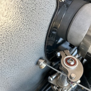 May include: Close-up of an engine component featuring a silver-colored throttle linkage and a coiled spring. The assembly is set against a textured, gray background, with a black component visible.