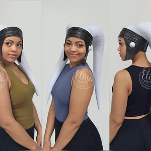 May include: A white and black headpiece with long, curved appendages. The headpiece has a black band and a silver button detail. The image shows the headpiece from multiple angles, showcasing its design and fit.