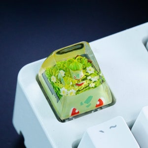 以下が含まれることがあります： 緑と黄色のキーキャップで、中に小さな詳細な人形が入っています。キーキャップは白いキーボードの上にあります。