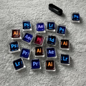 May include: A collection of square, clear resin keycaps with various Adobe software abbreviations in blue, orange, and red. Keycaps include "Ps", "Ai", "Ae", "Lr", "Pr", "Pi", and "Sd". A black, rectangular object is also present.