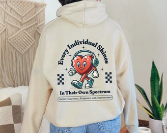 May include: A beige hoodie with a graphic print on the back. The graphic features a red heart with headphones, walking and smiling. The text reads "Every Individual Shines In Their Own Spectrum" with the words "Autism Awareness, Acceptance, and Empowerment" in smaller font below.