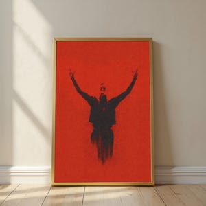 May include: Framed art print featuring a silhouette of a person with arms raised against a vibrant red background. The artwork is enclosed in a gold frame and leans against a neutral-toned wall, creating a striking contrast. The piece evokes a sense of freedom and expression.